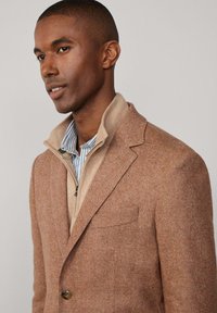 Blazer marrone a spina di pesce con colletto a fazzoletto, indossato sopra un maglione beige con zip e una camicia a righe blu. Texture liscia e vestibilità sartoriale.