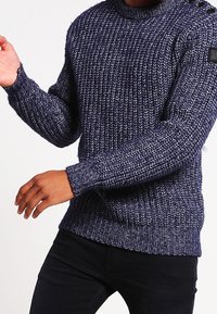 Navyblå stickad tröja med en strukturerad mönstring, långa ärmar och ribbade muddar. Har en rund halsringning och knappar på axeln.