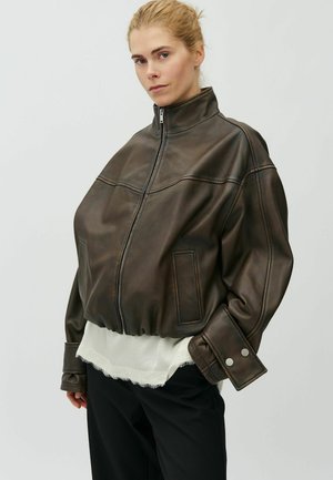 Jonge vrouw draagt een oversized donkerbruine leren jas met hoge kraag en rits, zwarte broek en wit shirt met rafelige zoom.