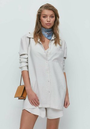 Junge Frau mit lockigem blonden Haar, die ein weißes, übergroßes Hemd, passende Shorts, ein blau gemustertes Halstuch und eine beige Schultertasche trägt.