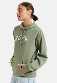 Green hoodie with white "ROXY" logo, drawstring hood, kangaroo pocket, and ribbed cuffs. Soft fabric and relaxed fit.