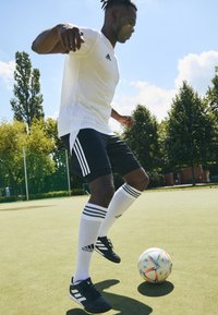 Vit Adidas t-shirt, svarta shorts med tre vita ränder och vita knälånga strumpor. Svarta fotbollsskor, dribblar en flerfärgad fotboll på grön konstgräs.