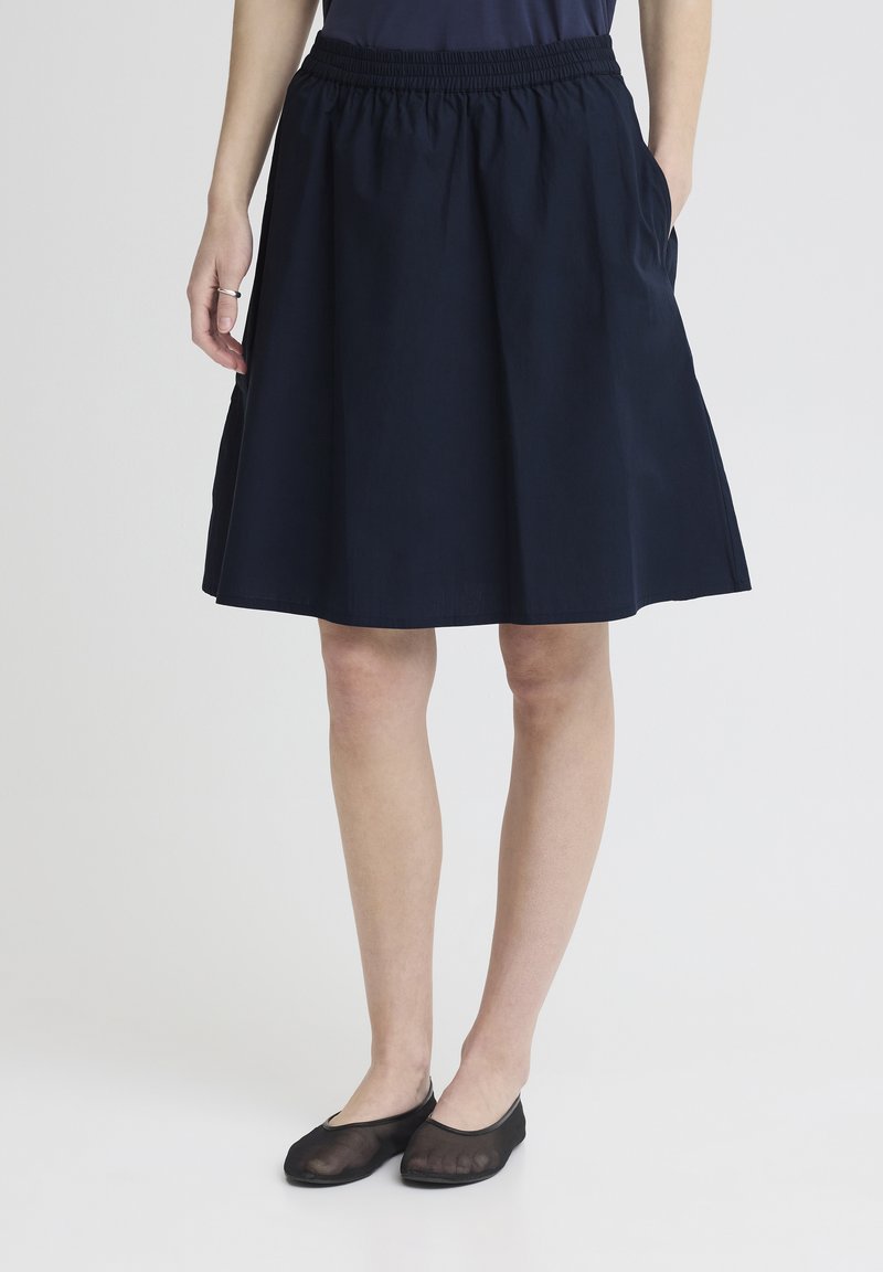 Femme portant une jupe bleu marine mi-longue avec une taille élastiquée et des ballerines noires transparentes, debout les mains dans les poches sur un fond uni.