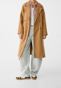 Manteau en mélange de laine marron avec une coupe croisée, des larges revers et des bandes contrastantes. Associé à un pantalon de survêtement gris et des baskets.
