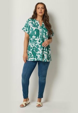 Mujer de pie contra la pared, vestida con blusa suelta de estampado paisley verde, jeans ajustados azules y sandalias blancas.