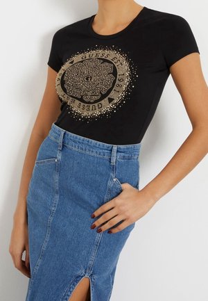 Woman wearing a black fitted t-shirt with gold circular floral design and text, paired with a high-waisted blue denim skirt with front slit.