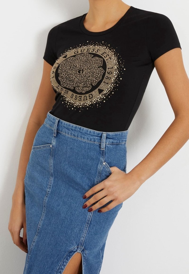 Femme portant un t-shirt noir ajusté avec un motif floral circulaire doré et du texte, associé à une jupe en denim bleu taille haute avec une fente à l'avant.