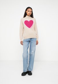 Beige gestrickter Pullover mit hohem Ausschnitt, der ein großes strukturiertes rosa Herzdesign auf der Vorderseite zeigt. Kombiniert mit hellblauen, zerrissenen Jeans und schwarzen Schuhen.