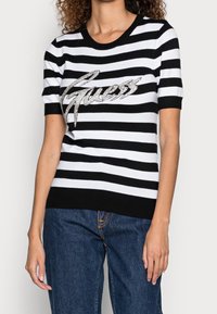 Czarnobiały, krótki sweter w paski z okrągłym dekoltem i błyszczącym logo „Guess” na wysokości klatki piersiowej. Tkanina o fakturze.