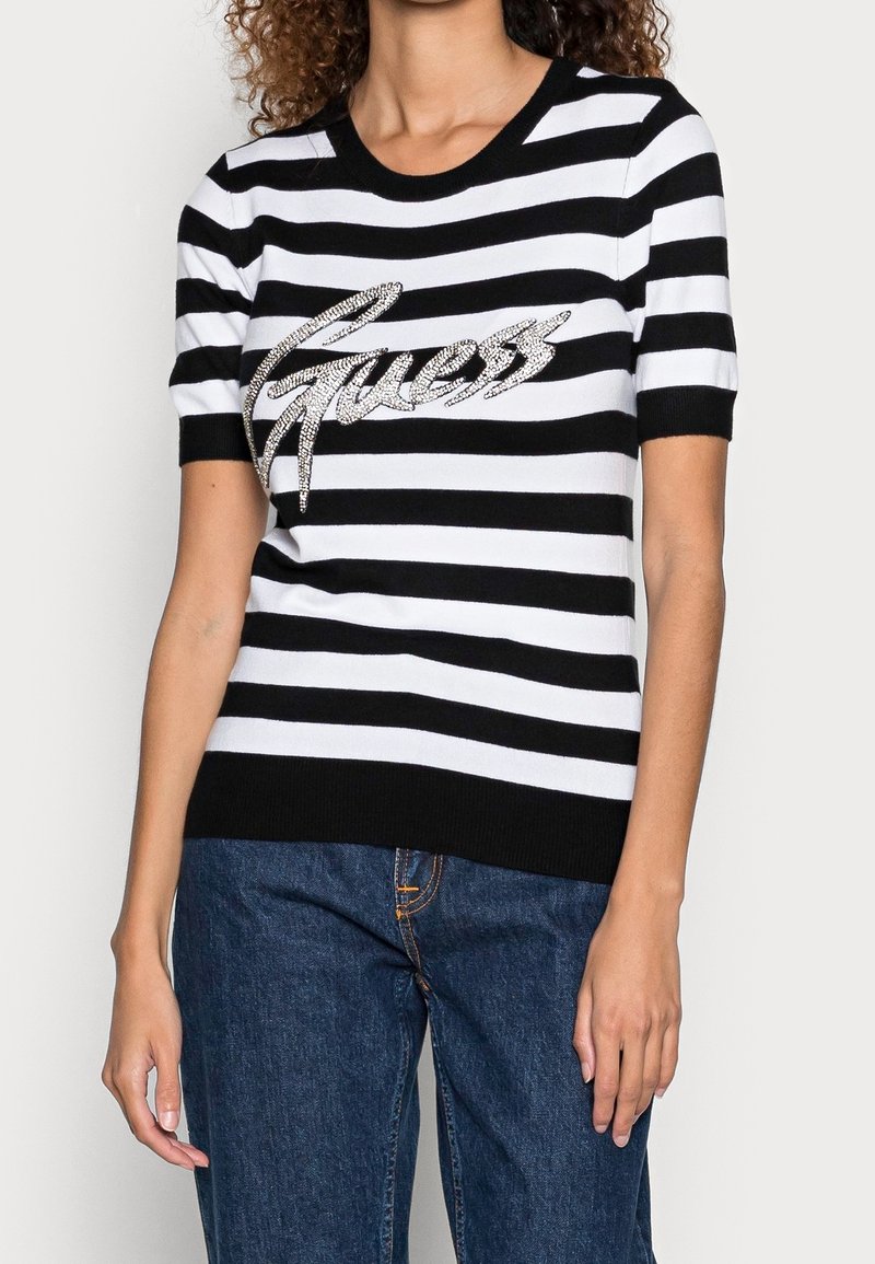 Czarnobiały, krótki sweter w paski z okrągłym dekoltem i błyszczącym logo „Guess” na wysokości klatki piersiowej. Tkanina o fakturze.