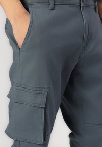 Zbliżenie na ciemnoszare szorty cargo z ręką częściowo włożoną w przednią kieszeń, podkreślające szwy i klapę kieszeni bocznej.