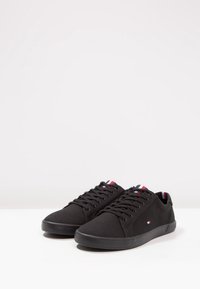 Schwarze Segeltuchschuhe mit flachen schwarzen Schnürsenkeln, Gummisohlen und kontrastierenden roten und blauen Akzenten auf der Zunge. Schlichte, strukturierte Oberfläche.