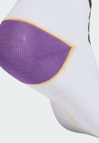 Biela ponožka s vystuženou fialovou pätou a tenkým žltým detailom na svetlom pozadí, ukazujúca pletenú textúru.