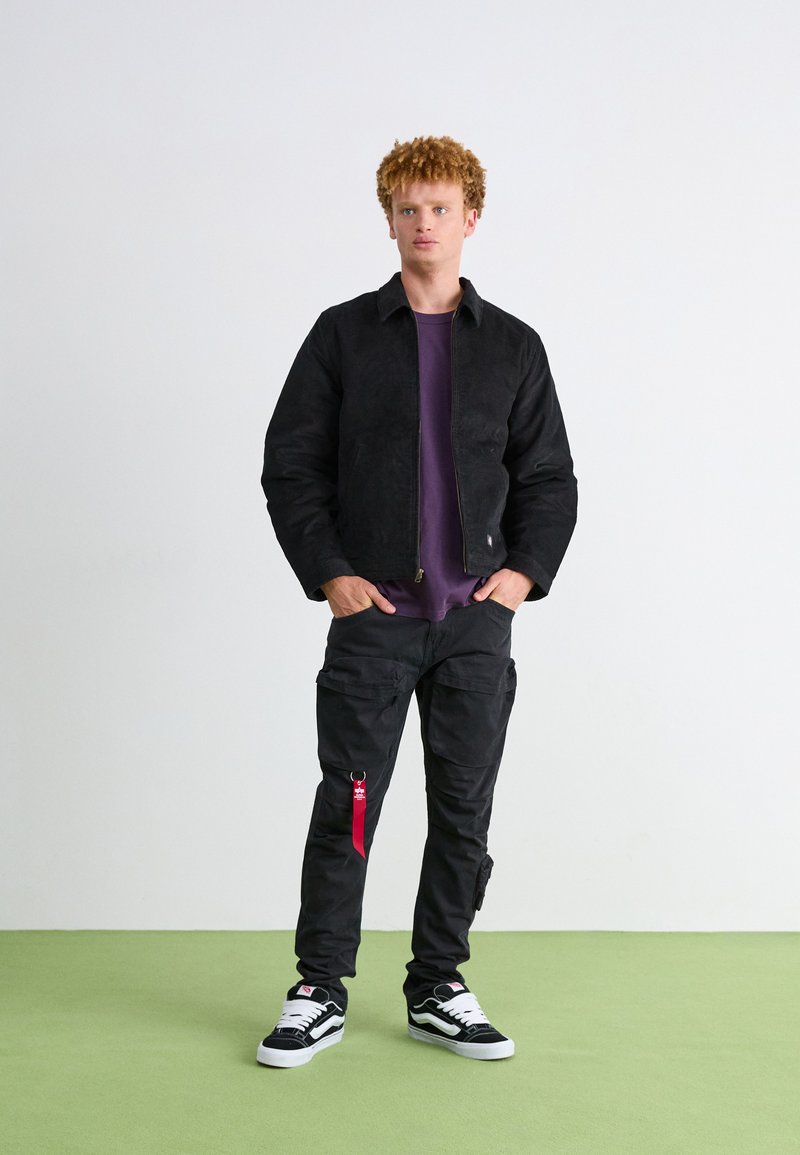 Czarna zamszowa kurtka, fioletowa koszula, czarne spodnie cargo z czerwonym brelokiem i czarne sneakersy z białymi akcentami na tle zielonej podłogi.