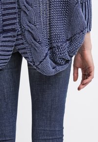 Suéter de punto ligero en azul con patrones texturizados, incluyendo trenzas y rayas, combinado con jeans de mezclilla oscuros.