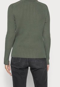Grüner gerippter Pullover mit langen Ärmeln, taillierter Silhouette, Rundhalsausschnitt und subtiler Textur, kombiniert mit dunkelgrauen Jeans.