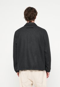 Sötétszürke gyapjú dzseki laza fazonnal, gallérral, látható záródás nélkül, és sima textúrával. Hátul szellőzőnyílással díszítve.