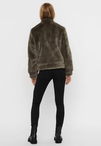 Kvinde iført en mørk olivengrøn faux fur-jakke, sorte skinny jeans og sorte ankelstøvler, stående med ryggen til mod en ensfarvet baggrund.
