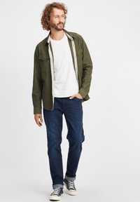 Chaqueta de camisas verde oliva sobre una camiseta blanca, combinada con jeans azul oscuro y zapatillas de deporte marinas. Presenta botones y un ajuste casual.