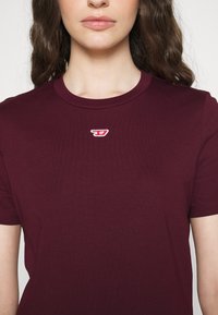 Burgundské tričko s kulatým výstřihem a krátkými rukávy, na hrudi s malým vyšitým logem v červené a bílé barvě.