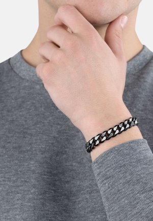 Bracciale intrecciato nero e argento su un polso, caratterizzato da anelli intrecciati e una finitura lucida, in contrasto con un maglione grigio.