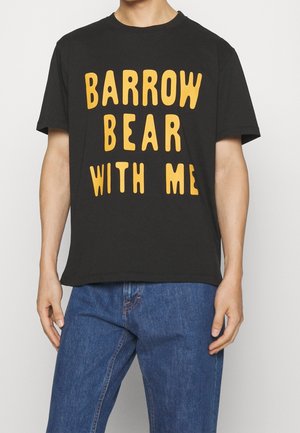 Maglietta nera con grande testo giallo "BARROW BEAR WITH ME" indossata da una persona in jeans blu, in piedi davanti a uno sfondo semplice.