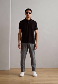 Schwarzes Polohemd mit rotem Logo, kombiniert mit hellgrauen Hosen und weißen Sneakers. Das Modell trägt eine dunkle Sonnenbrille, vor einem neutralen Hintergrund.