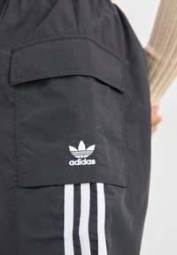 Czarne szorty cargo wykonane z lekkiego materiału, z kieszenią na klapie z przodu oraz białym logo Adidas nad trzema białymi paskami po bokach.