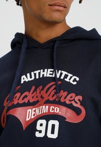 Sudadera azul marino con bolsillo canguro, que presenta un estampado gráfico en blanco y rojo que dice "Authentic Jack & Jones Denim Co. 90" en la parte frontal.