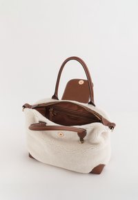 Bolso de mano de piel sintética de peluche en color crema texturizado con detalles en cuero marrón, doble asa y herrajes en tono dorado. El interior incluye un bolsillo con cremallera y compartimentos.