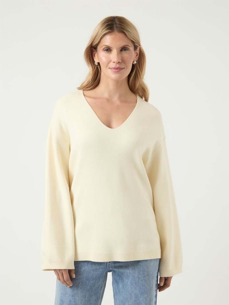 Pull en V color crème avec des manches longues et larges ; texture douce ; ourlet côtelé ; associé à un jean en denim bleu.