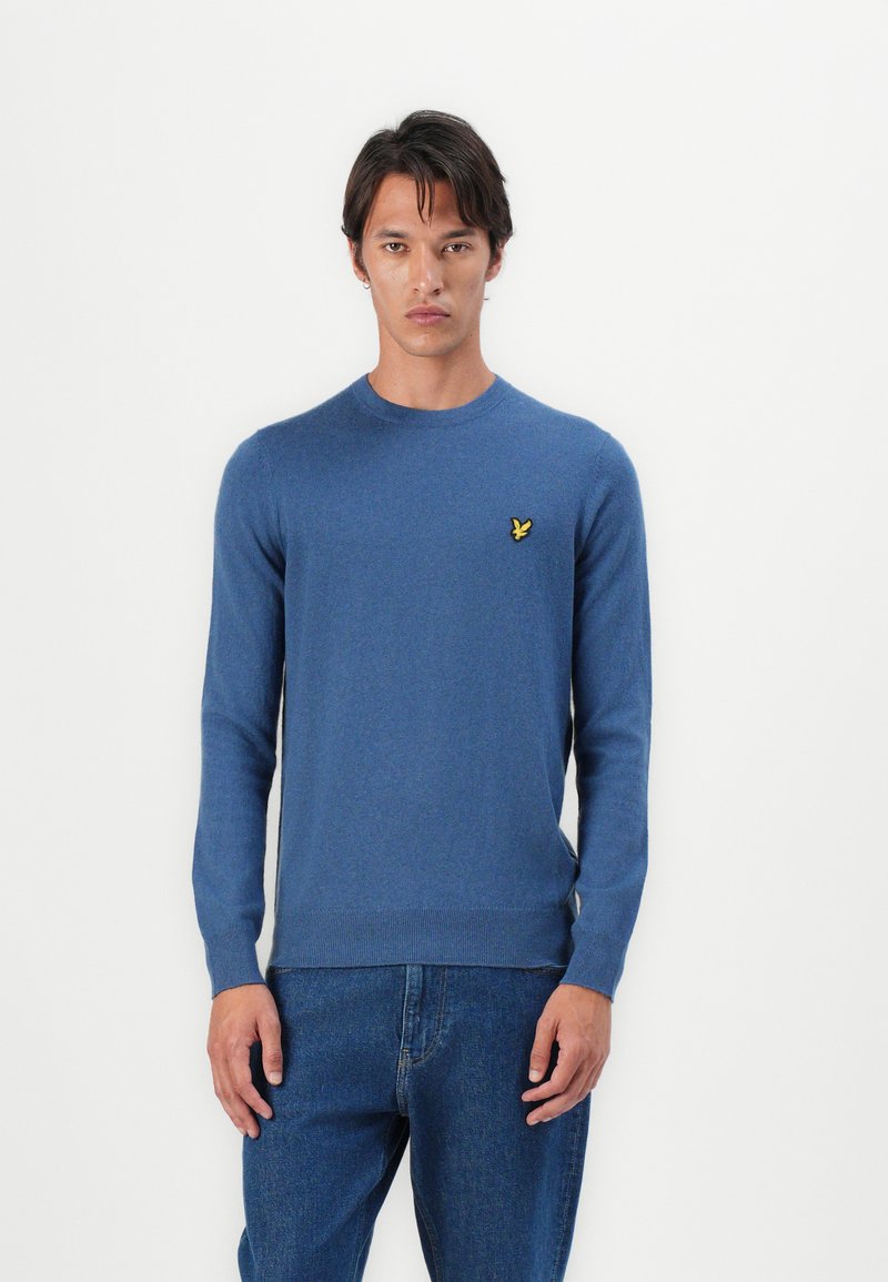 Pull à col rond bleu en tissu doux, avec un petit logo jaune brodé sur la poitrine gauche, des manches longues et des poignets côtelés.