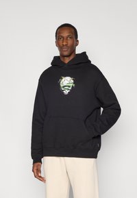 Μαύρο φούτερ με κουκούλα που φέρει γραφικό σχέδιο κρανίου με πράσινες λεπτομέρειες, κατασκευασμένο από μαλακό ύφασμα, με τσέπη καγκουρό και ριμπ μανσέτες.