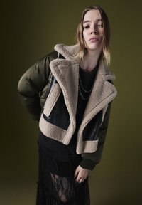 Junge Frau trägt eine zweifarbige Lammfelljacke, ein geschichtetes schwarzes Outfit mit einem Spitzenrock und silbernen Kettenhalsbändern, vor einem einfarbigen Olivgrünen Hintergrund.