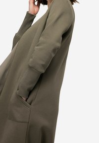 Oliven grøn lang cardigan, lavet af blødt stof, med ribbede manchetter og to sidelommer for funktionalitet.