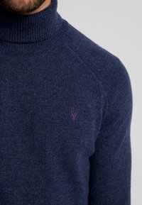 Pull à col roulé bleu marine avec col et épaules côtelés. Le matériau a une finition texturée. Présente un petit logo d'oiseau violet sur la poitrine.