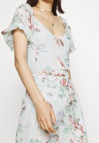 Robe florale en vert clair, avec des manches bouffantes, un décolleté en V, une taille ceinturée et une silhouette fluide agrémentée d'accents roses et bleus.