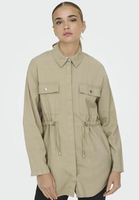 Bežová košeľa-jacket s dlhým rukávom, obojkovým golierom, sťahovacím pásom a dvoma náprsnými vreckami. Obsahuje strieborné cvoky a voľný strih.