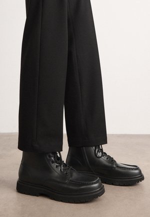 Schwarze Lederstiefel mit runden Zehen, sechs Ösen und strukturierten Gummisohlen, kombiniert mit maßgeschneiderten schwarzen Hosen.