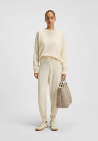 Helle beige Strickpullover und passende Jogginghose mit Kordelzug; gestepptes taupefarbenes Handtasche; cremefarbene Sneaker mit beiger Sohle.