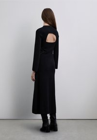 Langes schwarzes Strickkleid mit langen Ärmeln, einem Schlüssellochausschnitt auf der Rückseite und dekorativen Nieten an den Seiten. Kombiniert mit schwarzen Stiefeln mit Absatz.