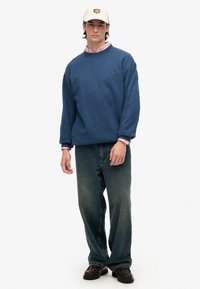 Sudadera azul de cuello redondo, de corte holgado, combinada con jeans de color azul claro. Gorro blanco con un emblema. Zapatos oscuros completan el look.