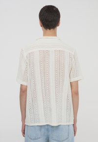Camicia bianca a maniche corte in tessuto a maglia aperta con strisce verticali, dotata di colletto e vestibilità rilassata.
