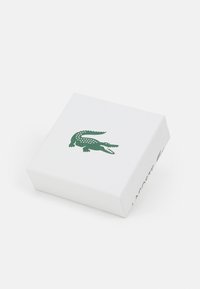 Bjela kutija s teksturiranim završetkom, s zelenim logotipom krokodila. Sa strane označeno "LACOSTE" u zelenoj boji. Kvadratnog oblika, minimalistički dizajn.