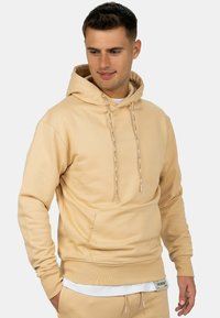 Uomo che indossa una felpa beige con cappuccio e pantaloni abbinati, guarda in basso e di lato con le mani rilassate.