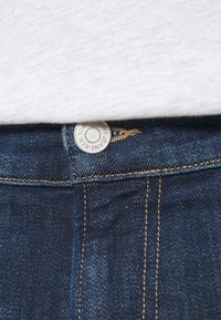 Tmavě modré džínové kalhoty s kovovým zapínáním na knoflík a zlatým stehem. Bílý látkový top částečně viditelný nad pasem.