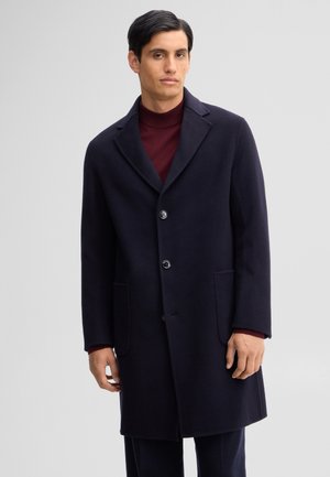 Uomo con capelli scuri che indossa un soprabito blu navy e un maglione dolcevita bordeaux su uno sfondo grigio chiaro uniforme.