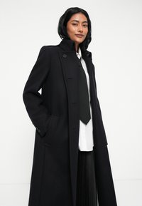 Cappotto lungo nero realizzato in tessuto testurizzato, con colletto alto, dettagli con bottoni e tasche laterali, indossato sopra una camicia bianca e una cravatta nera.