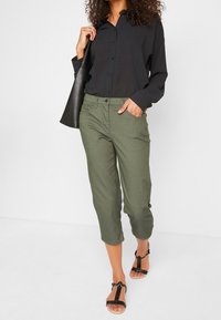 Pantalon court vert olive avec une finition texturée, associé à une chemise noire à manches longues et des sandales noires, avec un détail de poche élégant.