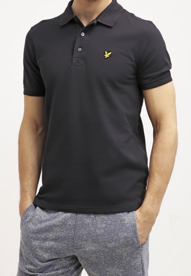 Homme portant un polo noir à manches courtes avec un logo d'oiseau jaune sur la poitrine et un pantalon décontracté gris, les mains dans les poches.